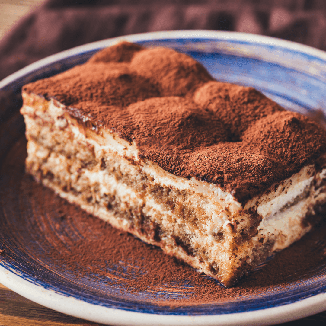 Close-up of a slice of tiramisu on a blue plate, topped with cocoa powder.