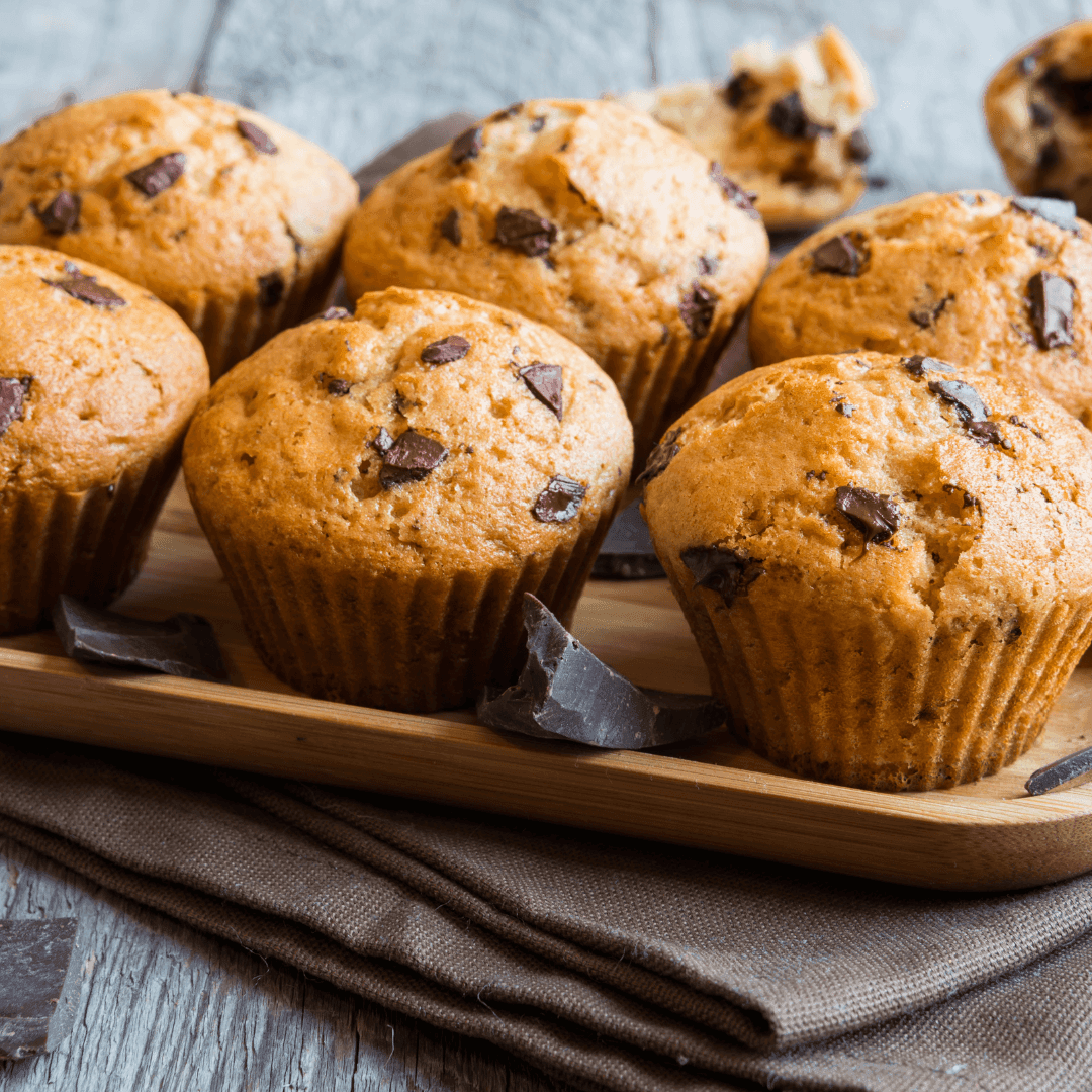 A wooden tray filled with freshly baked chocolate chip muffins arranged on a folded brown cloth.